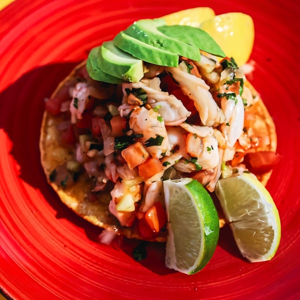 Loco Charlies' shrimp ceviche tostada with avocado slices, pico de gallo, and lime wedges on a red plate.