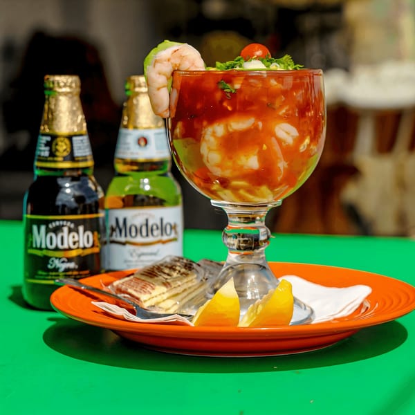 Shrimp cocktail in a goblet with Modelo beers, crackers, and lemon wedges on a green table.