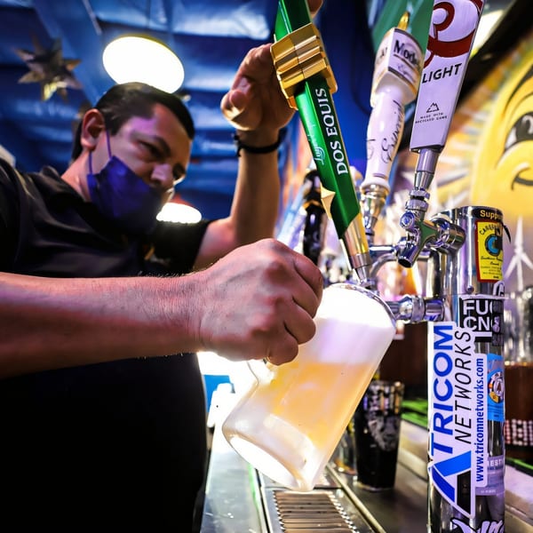 A bartender wearing a blue mask pours a Dos Equis beer from a tap into a glass pitcher.