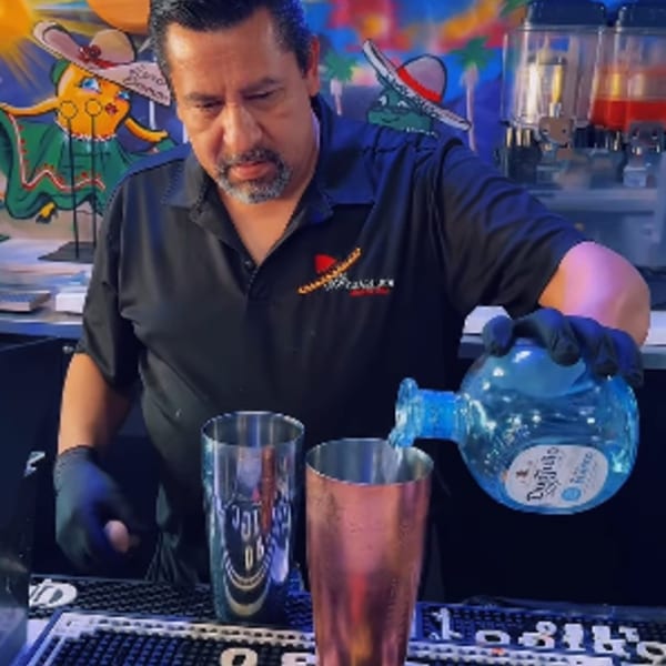 Bartender wearing black gloves pouring clear liquid from a blue bottle into a metal shaker.