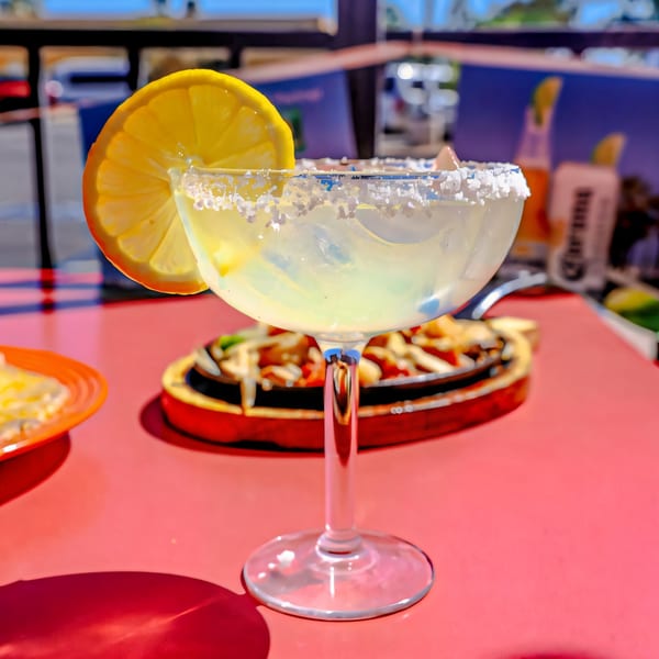Salt-rimmed margarita with a lemon slice on a red table, with fajitas in the background at Loco Charlies.